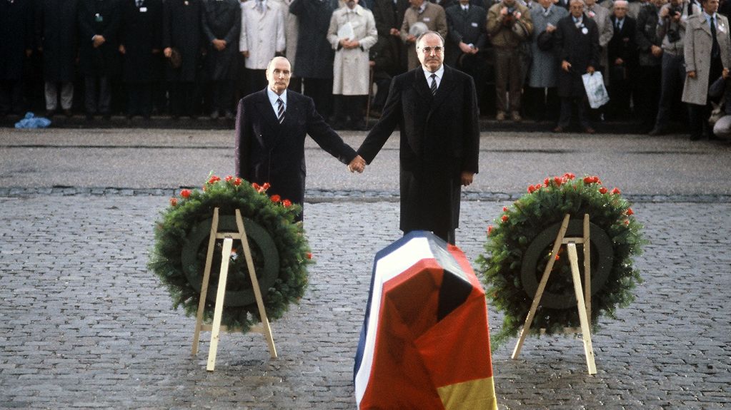Federal Chancellor Helmut Kohl and President François Mitterrand stand hand-in-hand before memorial wreaths.
