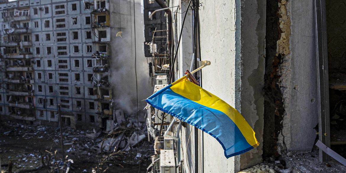 Eine Ukrainische Flagge weht am Fenster eines zerstörten Hauses in Donezk. Auch im Hintergrund ist ein zerstörter Wohnblock zu sehen.